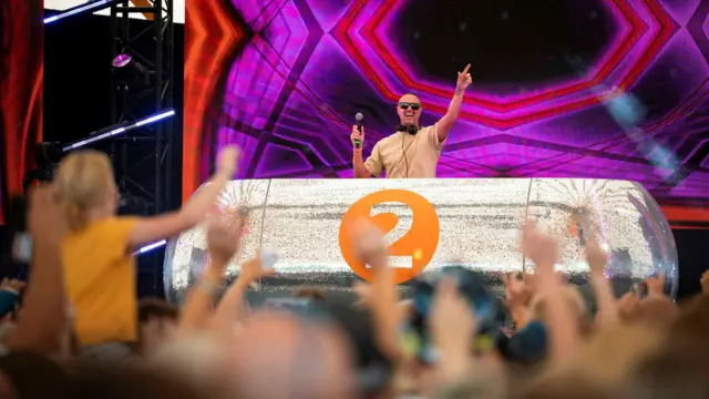 Paddy McGuinness raises his hand and holds a microphone on stage. He is behind a silver DJ desk and a crowd of people are dancing in front of him.