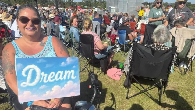 Anna Holmes is wearing a blue vest top, sunglasses and is sitting down around many other people. She is holding a blue sign that has clouds on it spelling out "dream".
