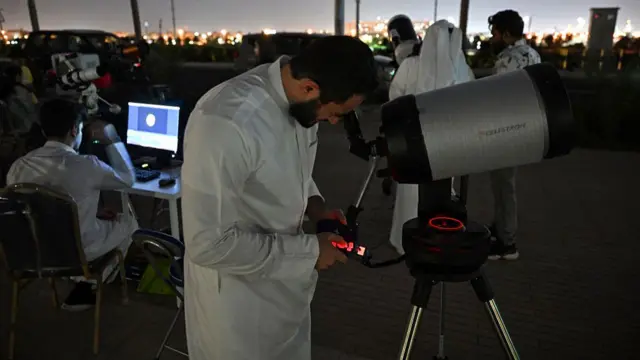 A man looking into the viewfinder of a telescope, another man is in the background looking at a picture of the moon onn a computer
