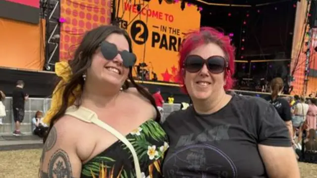 A woman with long dark hair wears a green floral dress and sunglasses in the shape of hearts. She stands next to another woman with red hair who is wearing sunglasses and a black T-shirt.