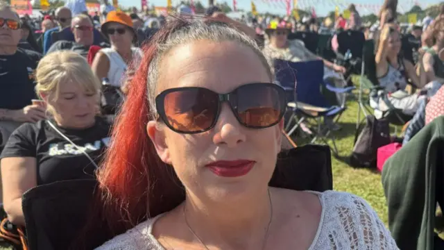 A woman wearing sunglasses and with red hair that is tied back sits in a camping chair. She smiles at the camera.
