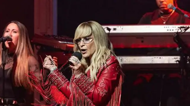 Anastacia singing on stage in front of a band. She has long blonde hair and glasses. She is wearing a red leather cowboy stale jacket with long red tassels along the arms. She is also wearing blue jeans and holding onto a microphone.