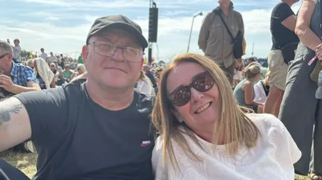A man wearing a black cap and T-shirt sits on the ground with a woman with shoulder length red hair and a white T-shirt.