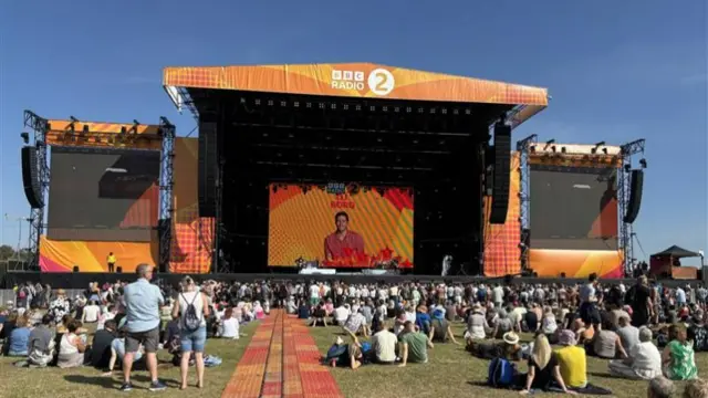A large main stage with BBC Radio 2 written on the top of it. A large screen sits in the middle of the stage. People attending the festival sit and stand in front of it.