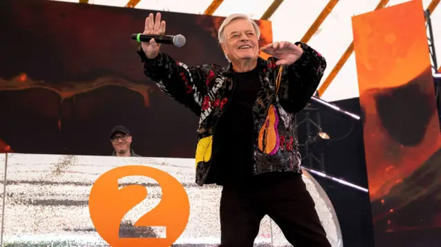 Tony Blackburn is wearing a black jacket with a guitar on it. He is dancing while holding a microphone on stage.