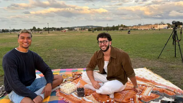 Jubilee and Daniel sitting on two picnic blankets, they have snacks including popcorn and grapes in front of them and there is a telescope visible in the background
