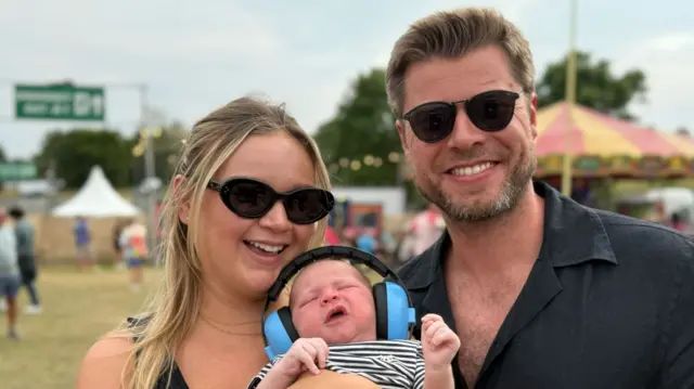 A woman and man smile at the camera with their baby held in their arms between them. The baby has blue ear defenders on. Both parents wear sunglasses. The woman has long blonde hair and the man has short brown hair and a beard.