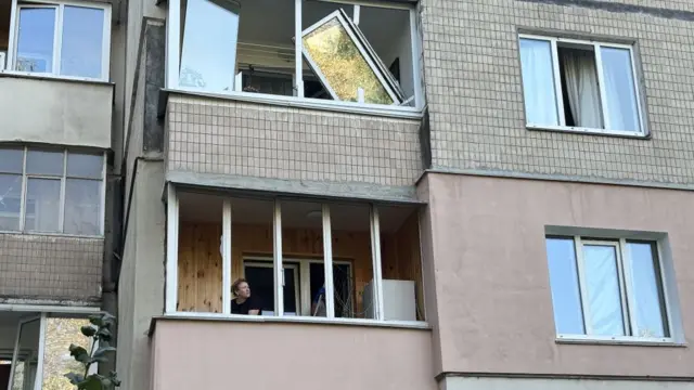 Two flats, one of which has had the windows blown. In the flat below, a person sits looking out