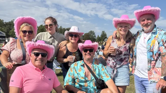 A group of people, some sitting, some standing, smile at the camera. Some wear pink cowboy hats and sunglasses.