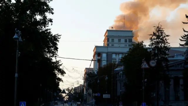 A tree-lined street with plumes of orange smoke rising above a building
