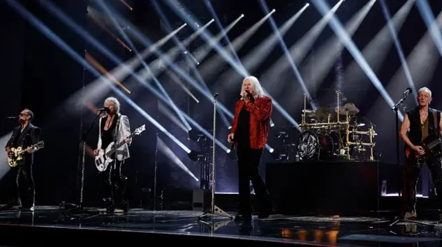 Def Leppard perform on stage. The band consists of five members four of which all stand behind microphone stands while another is behind a set of drums. Three of them play guitars while one only sings.