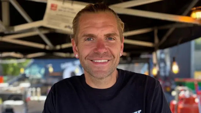 A man with blonde hair that is tied back behind his head smiles at the camera. He hate white circular earrings in and wears a black T-shirt as he stands underneath a gazebo.