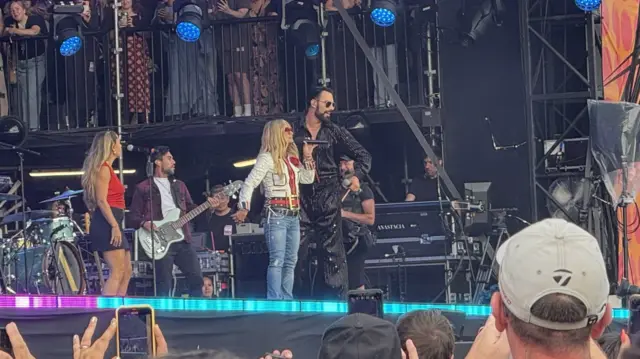 Rylan and Anastacia sing on stage together. They both hold microphones and are surrounded by band members and backing singers.