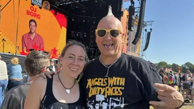 A woman and man smile at the camera. The man has one arm around the woman and his other hand is doing a rock hand sign. The man has a mohawk hairstyle and wears a black T-shirt. The woman has dark hair that has been tied back and she wears a black vest top.