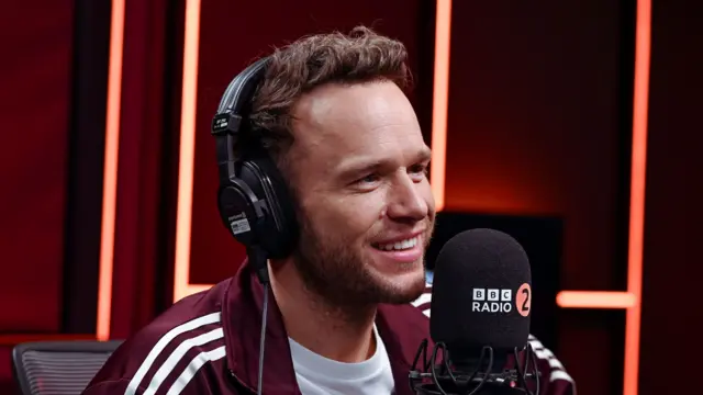 Olly Murs sits in a radio studio with a large microphone in front of him. He wears big headphones and a burgundy jacket with a white T-shirt underneath.