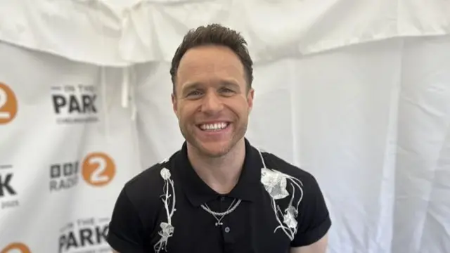 Olly Murs smiles at the camera. He has short dark hair, stubble and wears a black shirt with some white floral patterns on it.