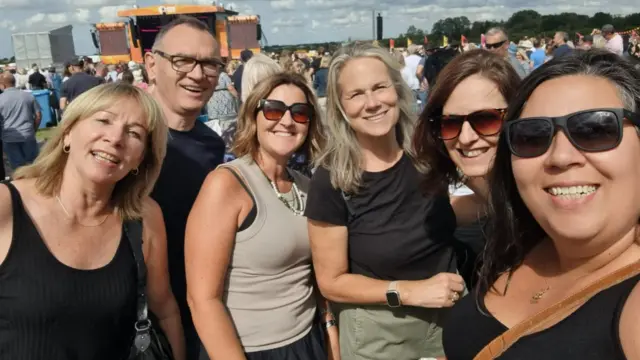 A group selfie of Penny Marshall, Martin Foster, Natasha Moshtagh, Sandra Ledwith, Claire Nicolls and Lisa Waters. They are all wearing black except for Natasha. They are smiling and have the sun shining on them. Behind them is the large festival crowd.