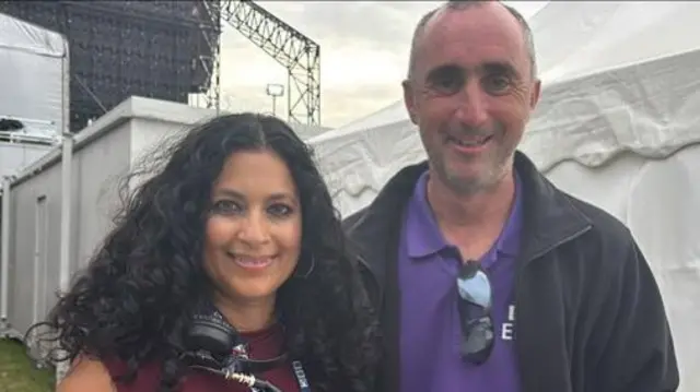 A woman with curly dark hair smiles at the camera. She has headphones around her neck. She stands next to a man wearing a purple polo top and blue jacket.