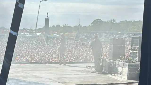 Sophie Ellis-Bextor is wearing a white mini skirt and heels on stage. Behind her is a man playing a guitar. In front is a large crowd watching her.