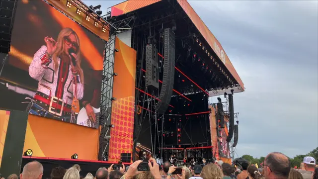 A view of a large festival stage. The crowd looks toward the singer who is performing. She can be seen on a large screen next to the stage.