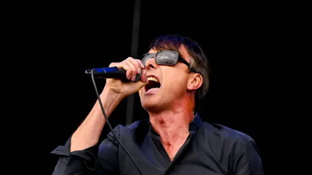 Suede frontman sings into a microphone. He has dark hair, sunglasses and a dark shirt.