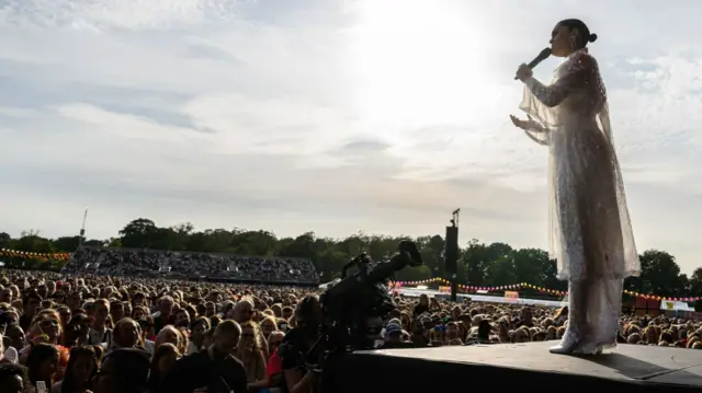 Jessie J wearing a white dress while singing on stage. Thousands of people watch on in front of her.