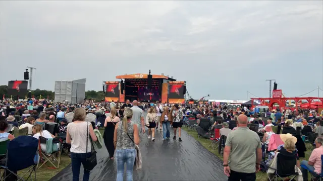 Hundreds of people watch a large screen in the distance, sitting in camping chairs, a walkway runs through the middle