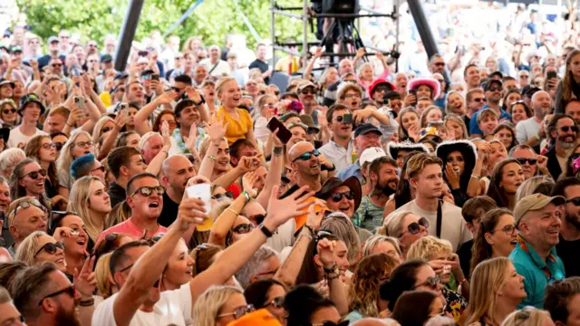 A general view of a busy crowd of people. Some wear hats, others have their arms in the air. Most are singing and smiling.