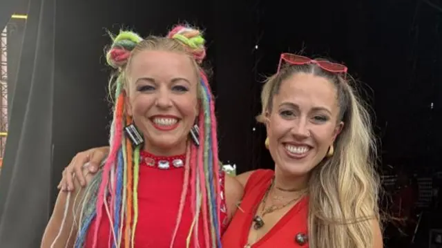 Two women wearing red tops have their arms around one another and smile at the camera. One woman has long blonde hair and the other has multicoloured dreadlocks.