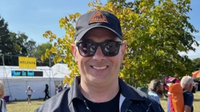 A man smiles at the camera wearing a cap with the Def Leppard band logo on it. He also has sunglasses on and a black shirt with a black T-shirt underneath.