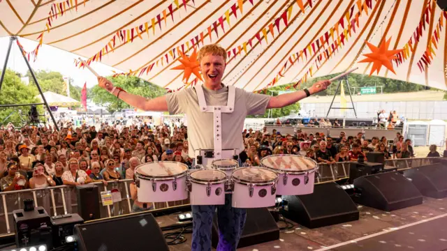 A man with red hair has a large set of drums strapped to his torso. He stands on a stage with the crowd behind him. He holds his arms out in the air as he takes the photo.