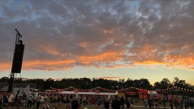 The sun sets over a festival. The clouds in the sky glow orange. Festival tents, lights and speakers as well as people can be seen milling around.