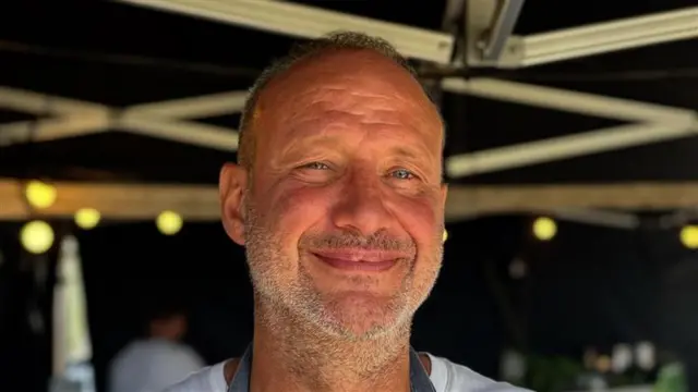 A man with short grey hair and grey stubble smiles at the camera underneath a gazebo. He wears a white T-shirt and chef apron.