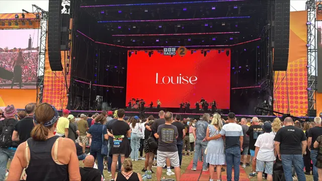 The name Louise is on a screen on a large stage. People watch on from the crowd.