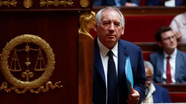 François Bayrou holding a blue folder walking to the lectern to speak in the National Assembly