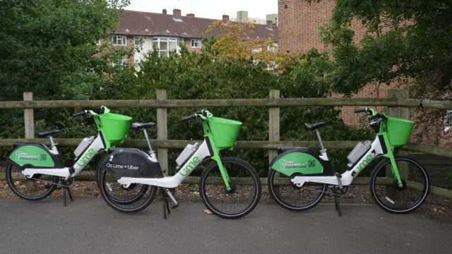 Three lime bikes parked in a row in front of a wooden fence. The bikes are white with black tyres and green front spokes and baskets.