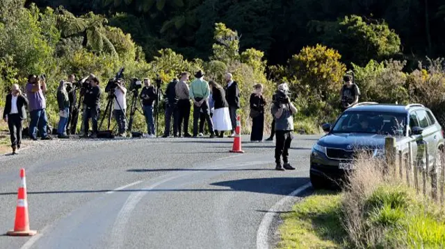 Members of the press gather along the side of a deserted road, with orange traffic cones cordoning off the road