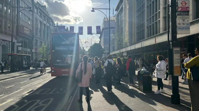 Bus drives past group of people waiting at bus stop on Oxford Street, London.