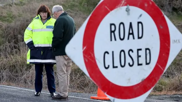 A white and red sign reading 'road closed' is in the foreground, a woman speaks to a man in the background