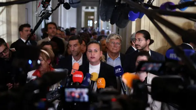 Mathilde Panot speaking in front of a crowd of reporters, holding microphones.