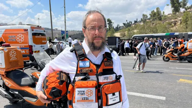A man looking at the camera wearing a bright orange emergency worker vest that has multiple pockets. He holds a helped, and there's a motorbike behind him