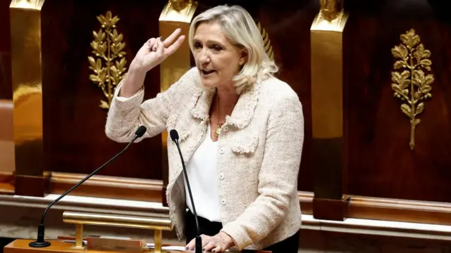 Marine Le Pen wears a cream jacket and gestures with her hand as she speaks behind a podium in the French National Assembly.