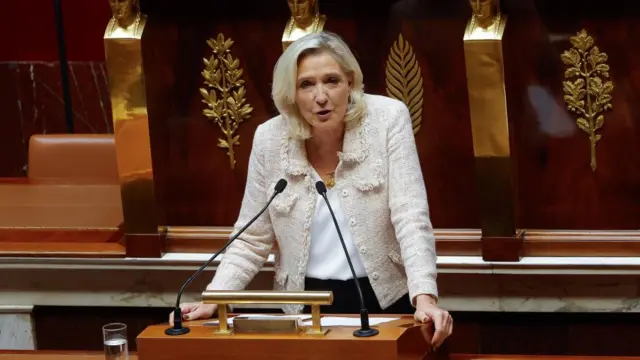 Marine Le Pen speaks behind a podium wearing a cream jacket in the National Assembly.