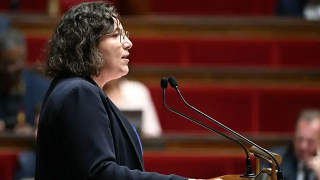 In a dark jacket and curly dark hair, a woman with glasses stands at a microphone
