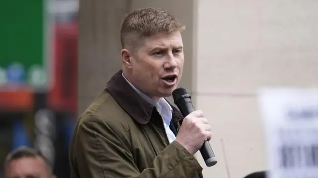 Eddie Dempsey wearing a green coat, speaking into a handheld wireless microphone.