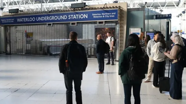 People standing on the concourse at Waterloo station in front of the entrance to the Underground station, which has a barrier stopping people getting in