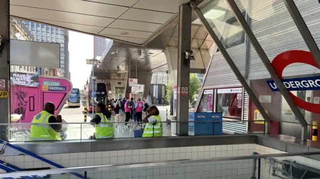 Three people wearing high vis jackets sitting along stairs with bus station behind them, large red double deckers in the background.