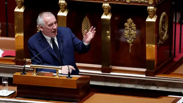 French Prime Minister Francois Bayrou delivers a statement in France's National Assembly. He gestures with his hand and speaks behind a podium, wearing a suit.