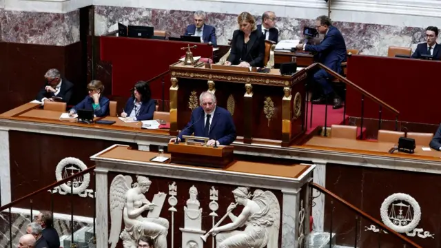French Prime Minister Francois Bayrou speaks in the National Assembly surrounded by other people. He speaks in a suit at a podium
