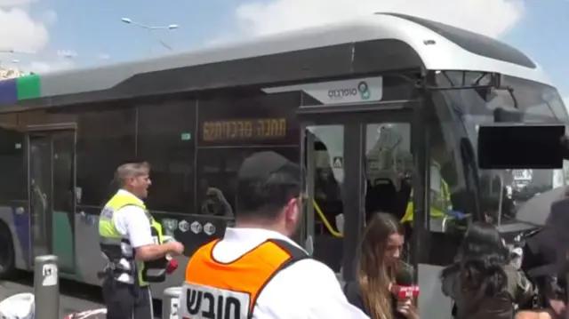 A bus being driven with smashed in doors as people watch.